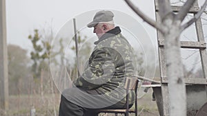 Portrait of concentrated senior Caucasian fisherman outdoors. Side view of old man sitting on chair on cloudy morning