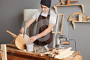Portrait of concentrated young adult man carpenter wearing white t-shirt, black cap and brown apron working in joinery, male doing