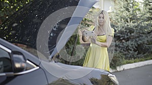 Portrait of concentrated young blondie taking photos of open car hood on smartphone. Young uncertain Caucasian woman in