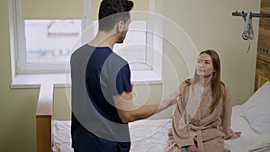 Portrait of concentrated young Caucasian woman sitting on hospital bed listening professional Middle Eastern man talking