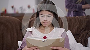 Portrait of concentrated young female student reading sitting on couch with blurred unrecognizable roommate walking at