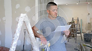 Portrait of a concentrated young man, standing near a stepladder and studying a document on a construction site.