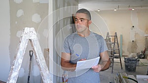 Portrait of a concentrated young man, standing near a stepladder and studying a document on a construction site.