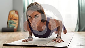 Portrait of concentrated young woman exercising, doing Four Limbed Staff yoga pose on a mat in living room at home