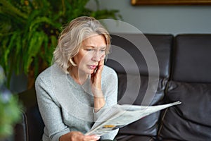 Portrait of a concerned woman reading bad news in a newspaper sitting on a couch in the living room at home.