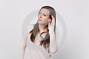 Portrait of concerned young woman in light clothes looking camera and eavesdrop with hearing gesture on white