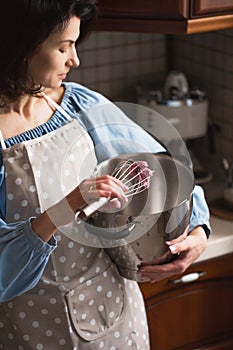 Portrait Of Confectioner Woman In Apron Preparing To Cook. Housewife is standing in the kitchen with a large pot and a whisk.