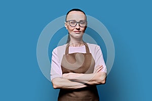 Portrait of confident 30s woman in apron on blue studio background