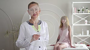 Portrait of confident acupuncturist posing with needles as young blond female patient sitting at the background. Adult