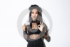 Portrait of confident asian woman assuring you in something, wearing halloween costume, showing okay gesture and credit
