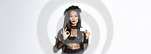Portrait of confident asian woman assuring you in something, wearing halloween costume, showing okay gesture and credit