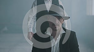 Portrait confident bad man sit on chair in fedora hat and other man walking from behind with a gun and shoot him in the