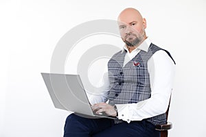Portrait of confident bald businessman in grey vest, blue jeans, white shirt, sitting on chair, holding laptop, posing.