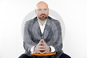 Portrait of confident bald middle-aged businessman in grey checkered jacket, vest, white shirt, sitting at stool posing.