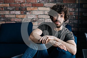 Portrait of confident bearded young man smiling looking at camera while sitting on comfortable sofa in cozy living room