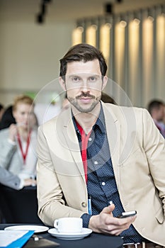 Portrait of confident businessman holding smart phone while sitting at convention center