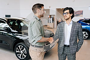 Portrait of confident car dealer in business suit welcoming young man customer in showroom. Professional salesman