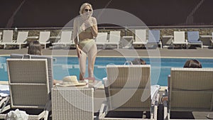 Portrait of confident Caucasian blond woman in sunglasses talking to friends lying on sunlounges in front of pool. Wide