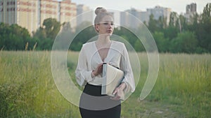 Portrait of confident Caucasian businesswoman standing in city park with documents folder and waiting. Young beautiful