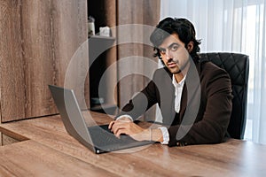 Portrait of confident CEO wearing suit working on laptop computer sitting at office desk. Successful businessman with