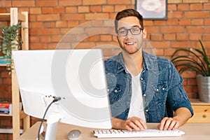 Portrait of confident cheerful handsome young freelance designer male in stylish glasses working on desktop computer