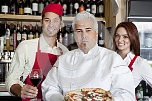 Portrait of a confident chef holding pizza with wait staff in background