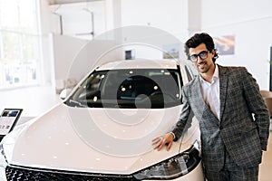 Portrait of confident customer male in business suit touching hood of new car standing looking at camera with positive