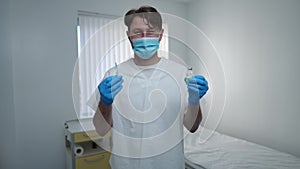 Portrait of confident doctor in face mask posing with syringe and ampoule of coronavirus vaccine in hospital ward