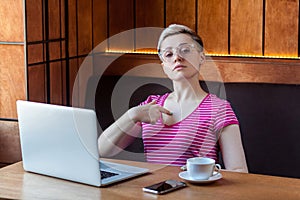 Portrait of confident egoist young girl with short hair in pink t-shirt and eyeglasses is sitting alone in cafe and pointing photo