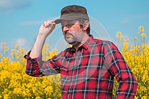 Portrait of confident farmer in blooming rapeseed field. Farm worker wearing red shirt and brown trucker's hat at plantation