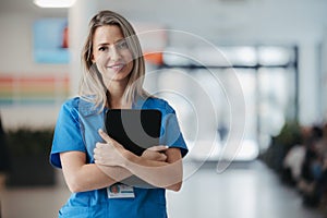 Portrait of confident female doctor in hospital corridor. Beautiful nurse wearing blue uniform, holding clipboard