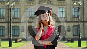 Portrait of confident girl in black mantle and cap standing in front of university building. Young woman celebrating her