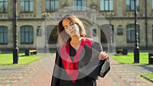 Portrait of confident girl in black and red mantle holding cap standing in front of university building. Young woman