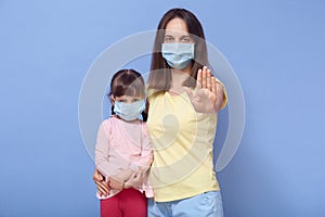 Portrait of confident good looking young female making gesture, raising hand, hugging her daughter, being in medical masks,