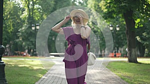 Portrait confident joyful slim woman in elegant purple jumpsuit and straw hat strolling in sunlight in summer park