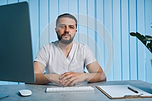 Portrait of confident male doctor sitting at desk at workplace and looking camera. Man physician