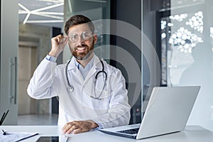 Portrait of a confident male doctor sitting at a workplace in a hospital, smiling at the camera while holding glasses
