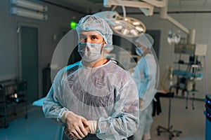 Portrait of confident male doctor in surgical mask and hat in operating room. Practitioner standing posing looking at