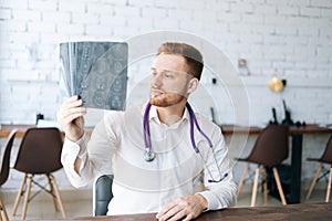 Portrait of confident male doctor wearing white uniform examining brain computerized tomography scan sitting at desk in