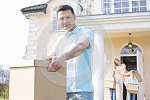 Portrait of confident man carrying cardboard box while moving house with family in background