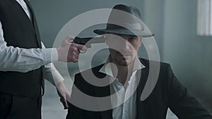 Portrait confident man sit on chair in fedora hat and other man walking from behind with a gun and shoot him in the head