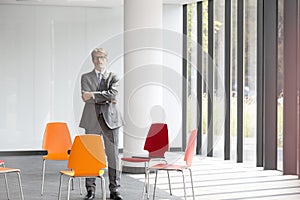 Portrait of confident mature businessman standing with arms crossed amidst chairs in new office