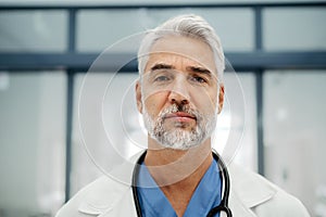 Portrait of confident mature doctor standing in Hospital corridor. Handsome doctor with gray hair wearing white coat