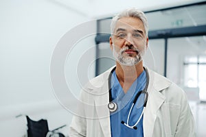 Portrait of confident mature doctor standing in Hospital corridor. Handsome doctor with gray hair wearing white coat