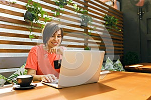 Portrait of confident mature professional woman in glasses, a coral T-shirt sitting on summer terrace in cafe, using laptop