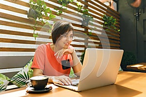 Portrait of confident mature professional woman in glasses, a coral T-shirt sitting on summer terrace in cafe, using laptop
