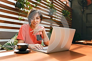 Portrait of confident mature professional woman in glasses, a coral T-shirt sitting on summer terrace in cafe, using laptop