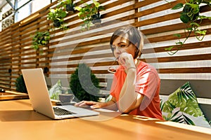 Portrait of confident mature professional woman in glasses, a coral T-shirt sitting on summer terrace in cafe, using laptop