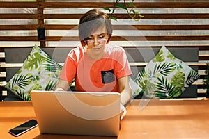 Portrait of confident mature professional woman in glasses, a coral T-shirt sitting on summer terrace in cafe, using laptop