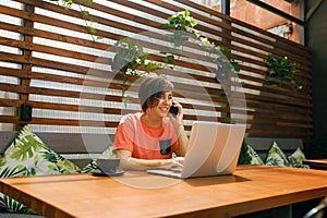 Portrait of confident mature professional woman in glasses, a coral T-shirt sitting on summer terrace in cafe, using laptop